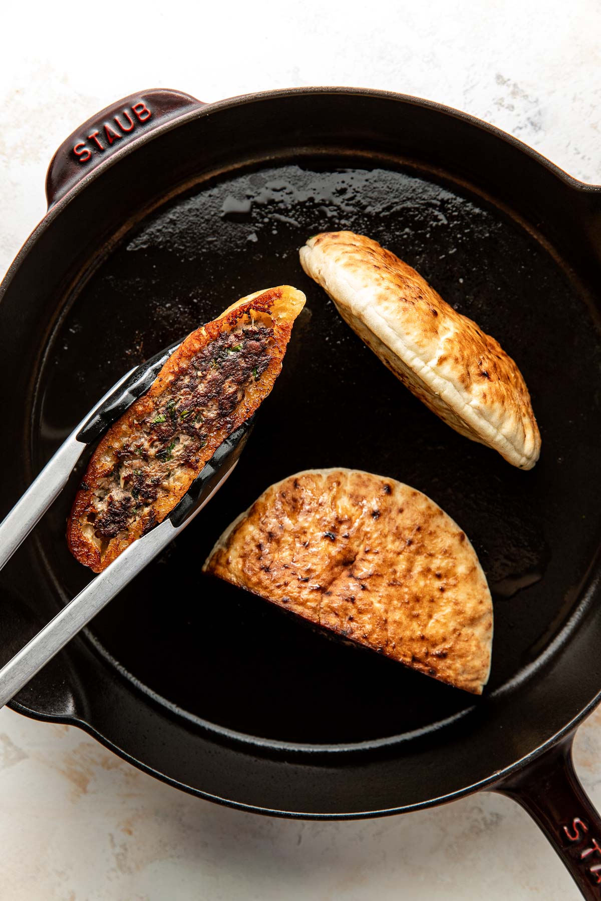 A skillet with two folded pita breads being cooked; one is held open with tongs to reveal a dark, seasoned filling inside.