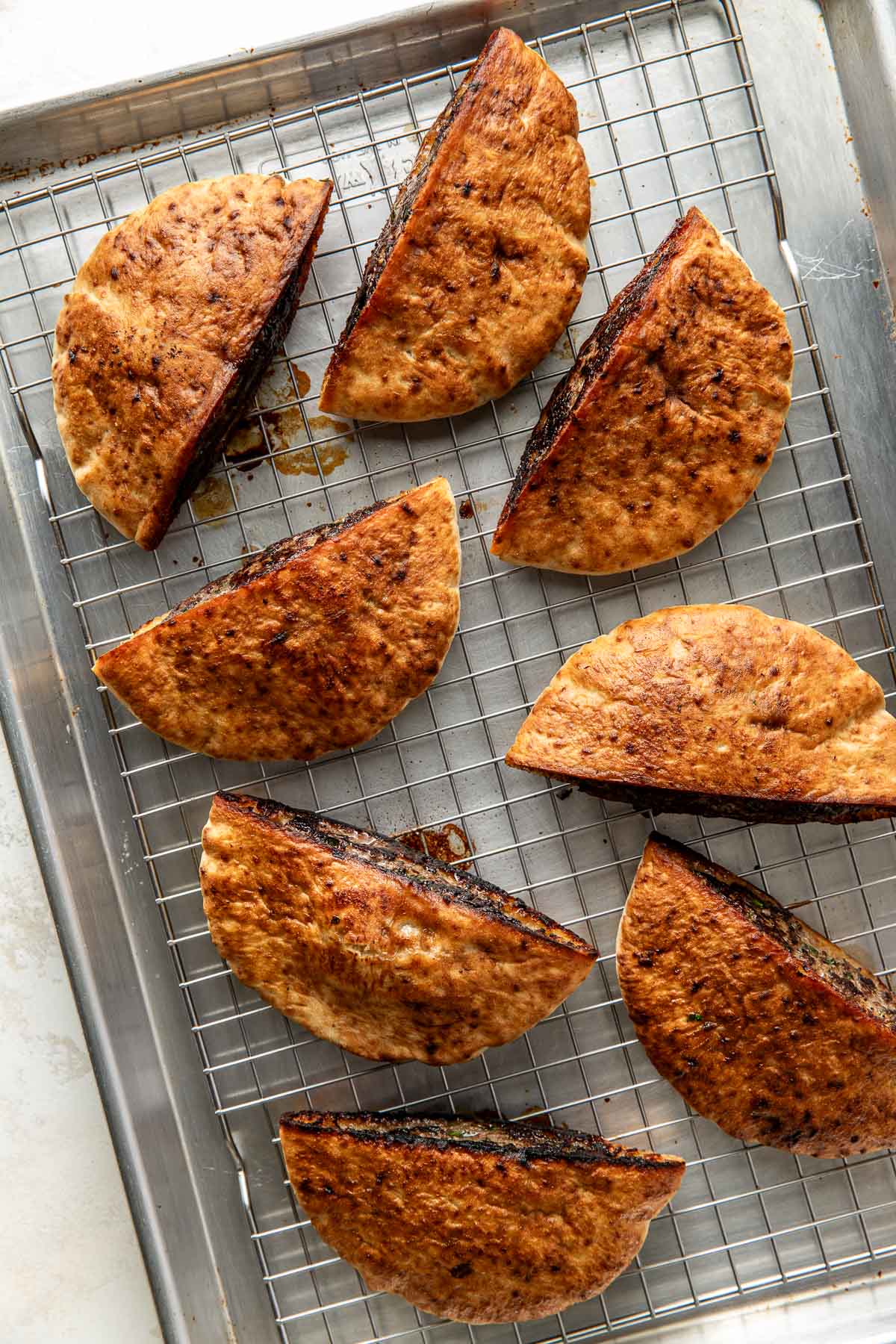 Eight folded pita breads filled with a dark filling are arranged on a wire rack set over a baking tray, suggesting they have been baked or toasted. The pitas have a golden-brown, crispy exterior.