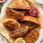 A white platter holds several pita bread halves stuffed with a dark spiced meat filling, garnished with chopped herbs, and served with lemon wedges, pickled onions, and bowls of salad and sauce on the side.