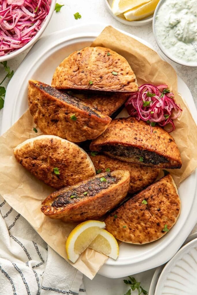 A white platter holds several pita bread halves stuffed with a dark spiced meat filling, garnished with chopped herbs, and served with lemon wedges, pickled onions, and bowls of salad and sauce on the side.
