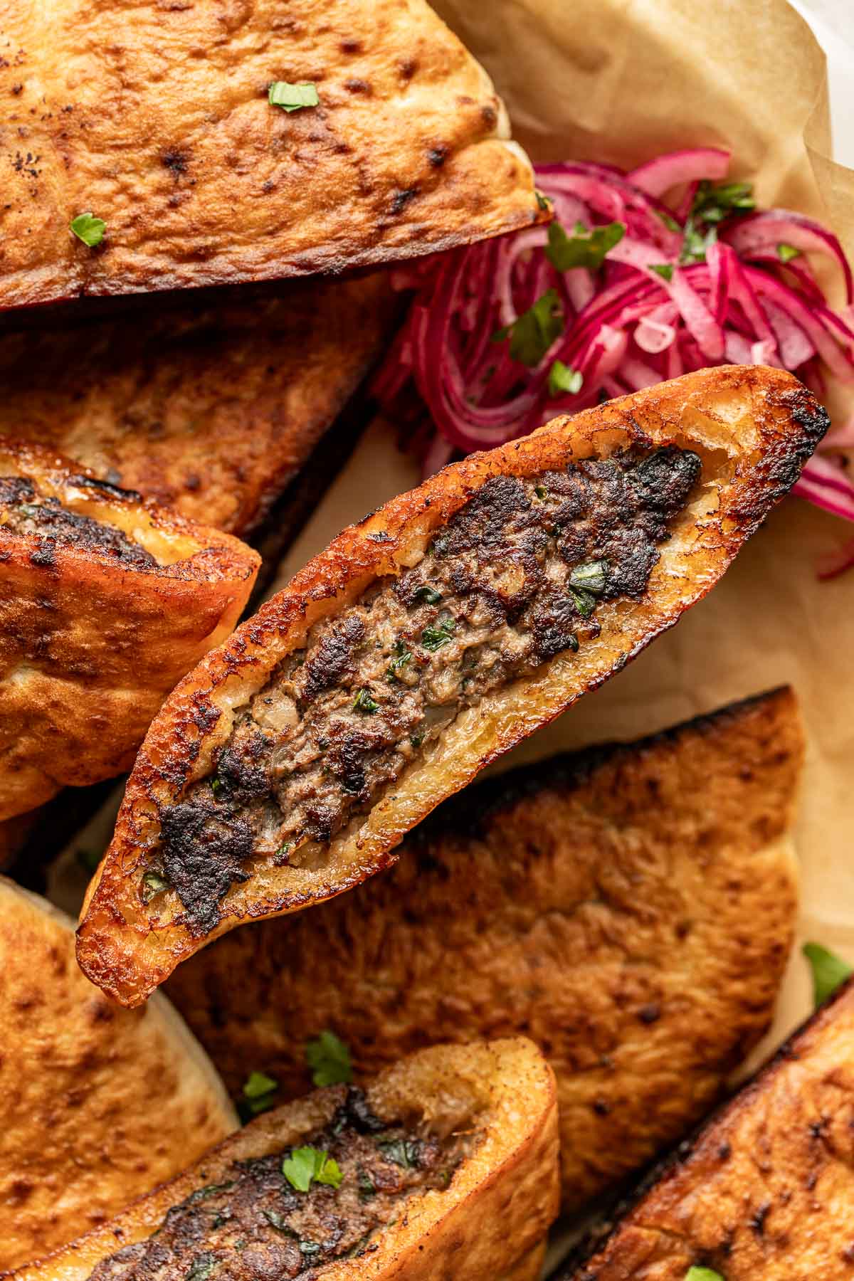 A close-up of baked Turkish pide bread topped with spiced ground meat, garnished with herbs, surrounded by more pide and pickled red onions on parchment paper.