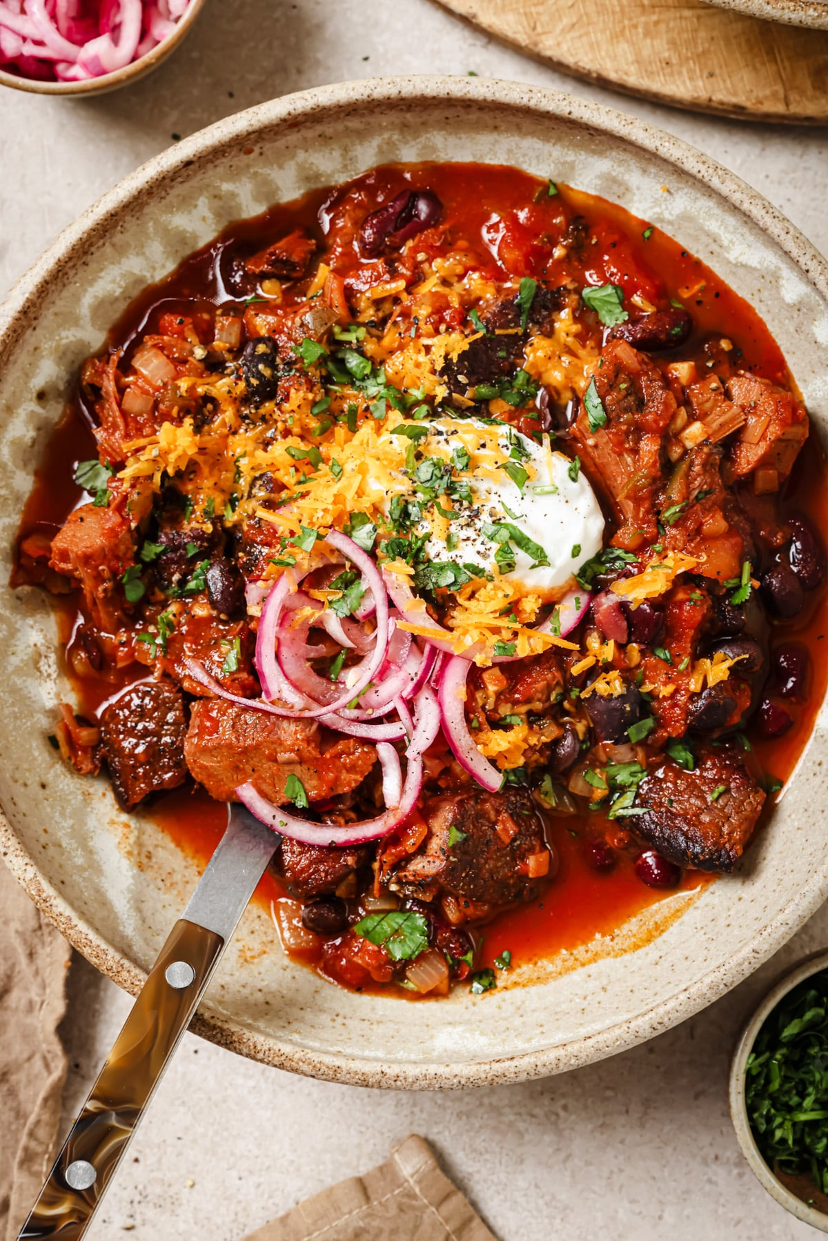A bowl of chili with chunks of beef, kidney beans, topped with shredded cheese, sour cream, sliced red onions, and chopped herbs. A spoon rests in the bowl, and small dishes sit nearby.