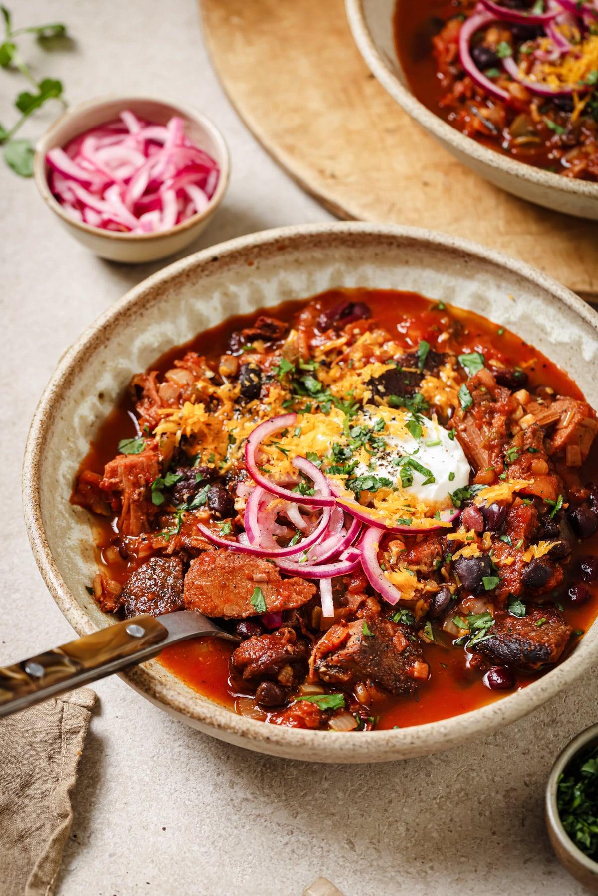 A bowl of chili topped with shredded cheese, sour cream, pickled red onions, and chopped herbs sits on a table with a knife resting on the bowl. Pickled onions and another bowl of chili are visible in the background.