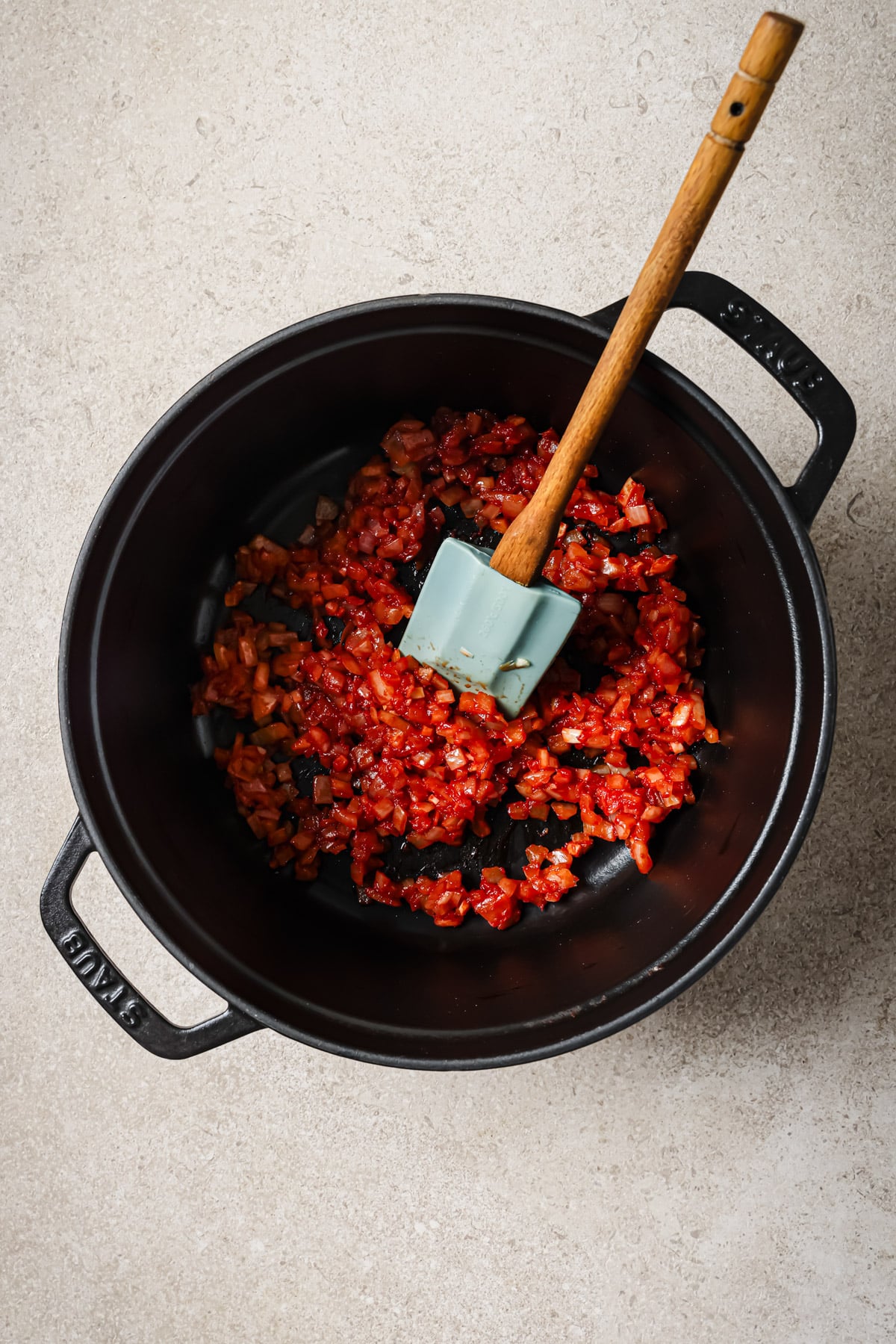 A black pot on a light surface containing sautéed chopped onions, tomatoes, and vegetables, with a blue spatula and wooden handle resting inside.