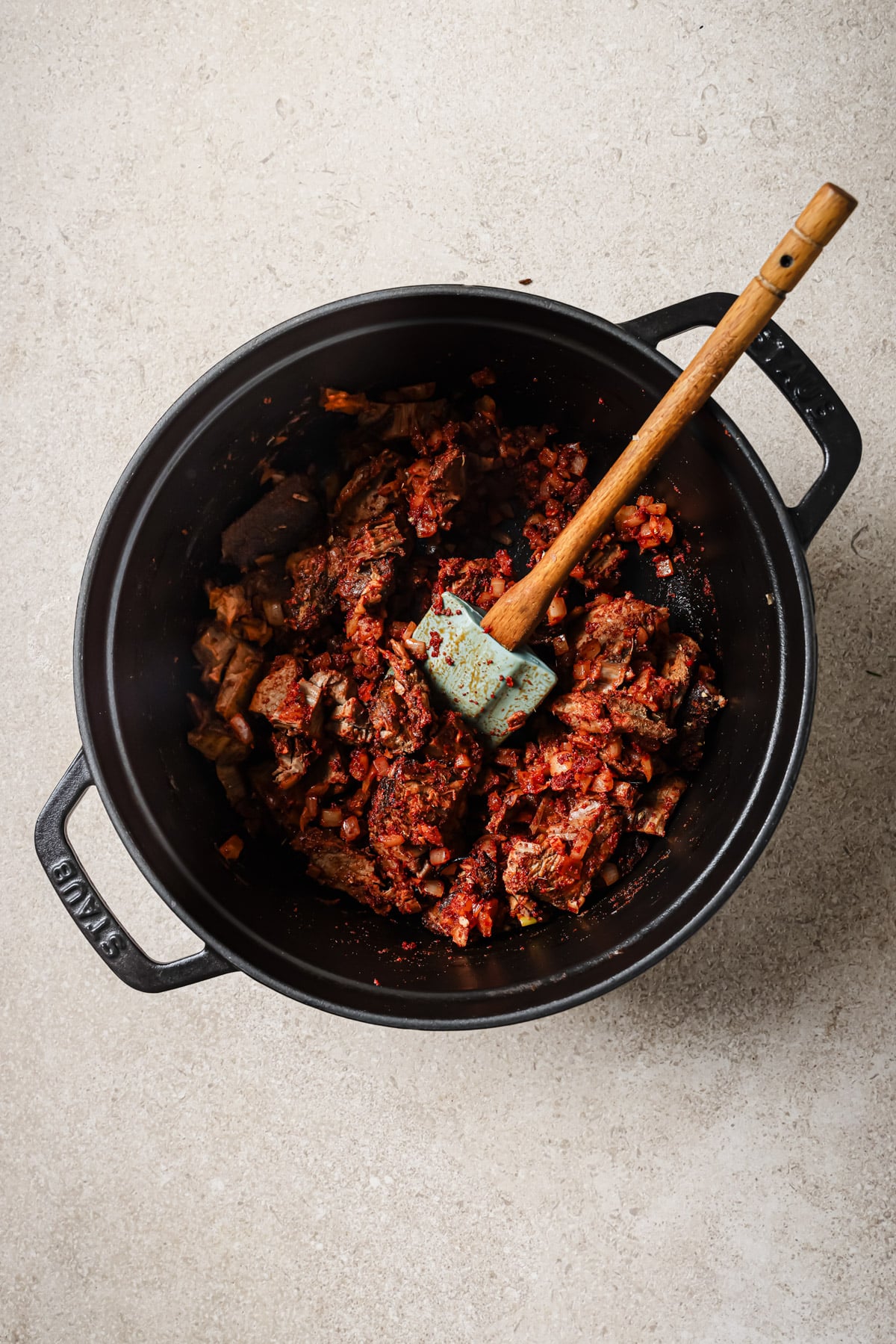 A black pot on a light countertop contains sliced beef and chopped onions mixed with red spices, being stirred with a wooden and green spatula.