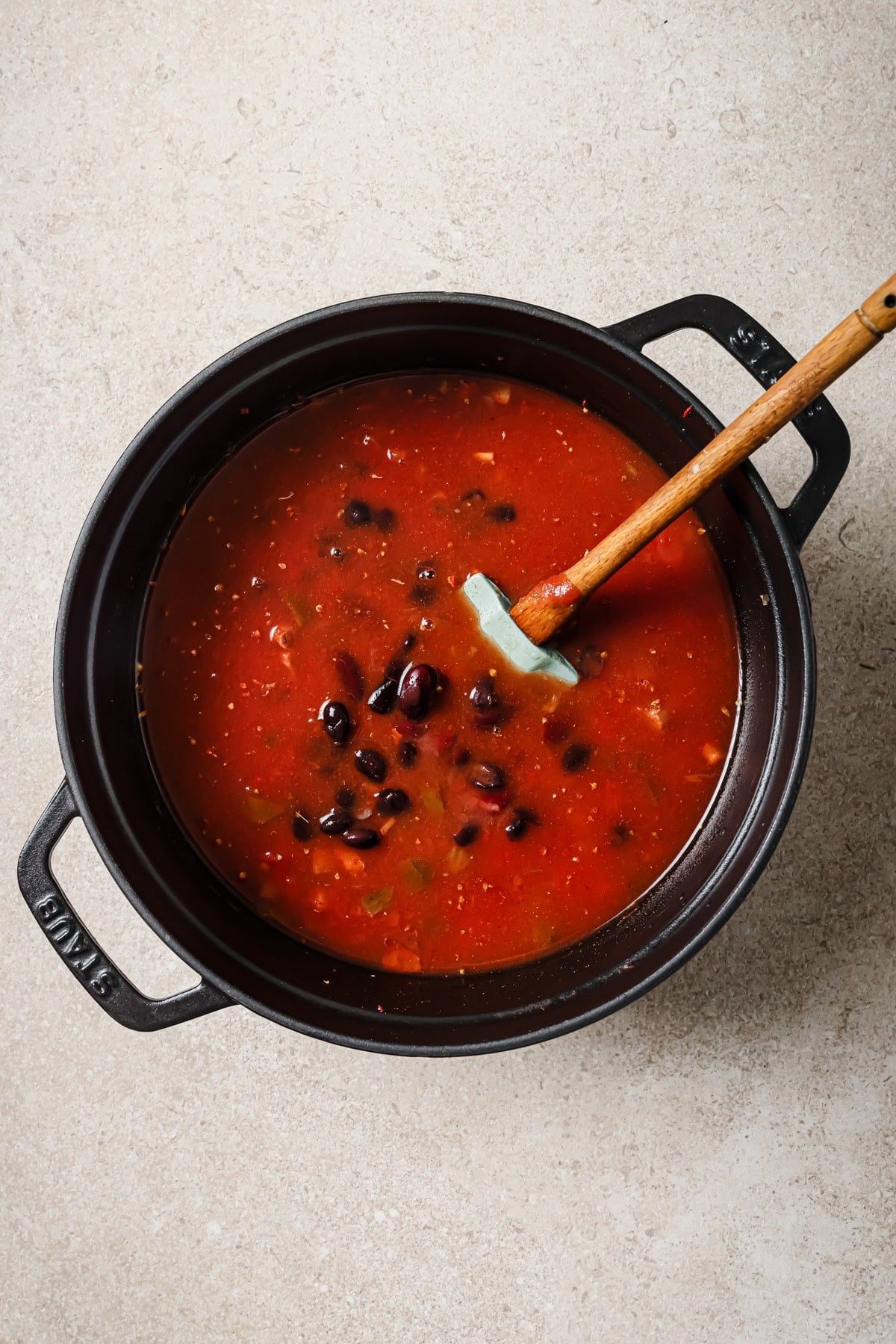 A black pot filled with red tomato-based soup containing beans and vegetables sits on a light countertop. A wooden spoon with a blue silicone tip rests inside the pot.