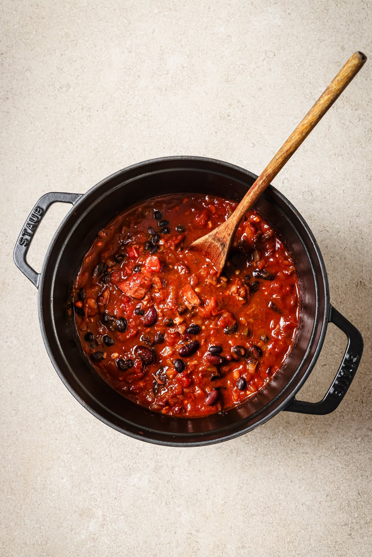 A black pot filled with chunky tomato and bean chili sits on a light countertop, with a wooden spoon resting inside the pot.