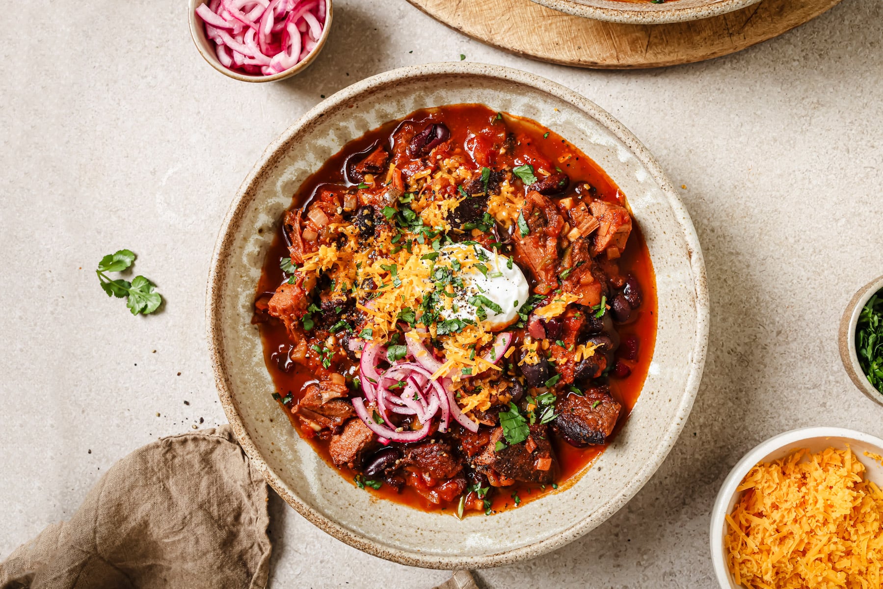 A bowl of chili topped with shredded cheese, sour cream, pickled red onions, and chopped cilantro. Surrounding the bowl are small dishes of shredded cheese, pickled onions, herbs, and a napkin.