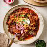 A bowl of chili topped with sour cream, shredded cheese, sliced red onions, and chopped herbs sits on a table with small bowls of pickled onions and fresh herbs nearby. A fork rests in the bowl.