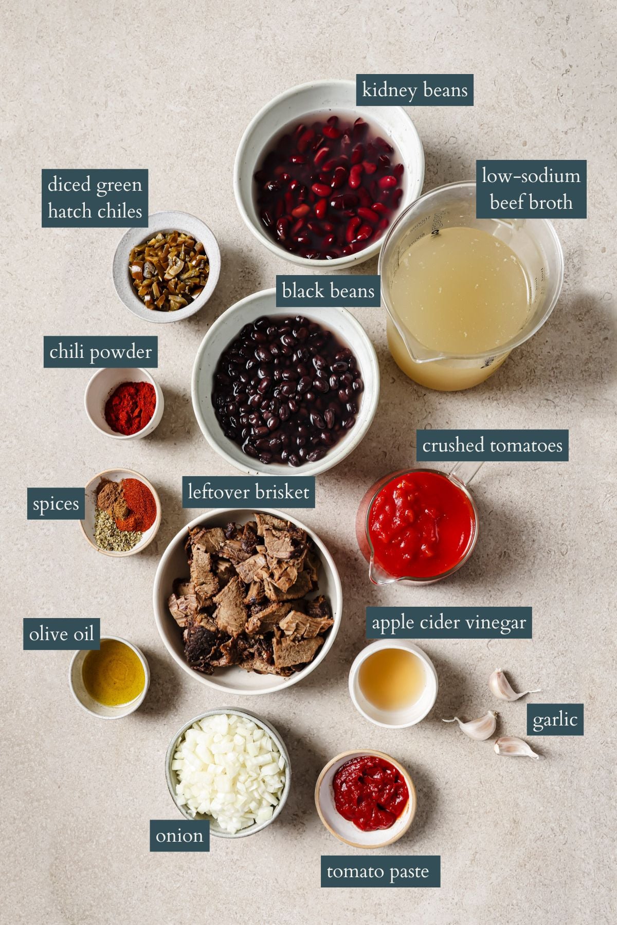 Overhead view of labeled chili ingredients in bowls and measuring cups: kidney beans, black beans, beef broth, crushed tomatoes, diced green chiles, chili powder, spices, leftover brisket, olive oil, onion, tomato paste, apple cider vinegar, and garlic.
