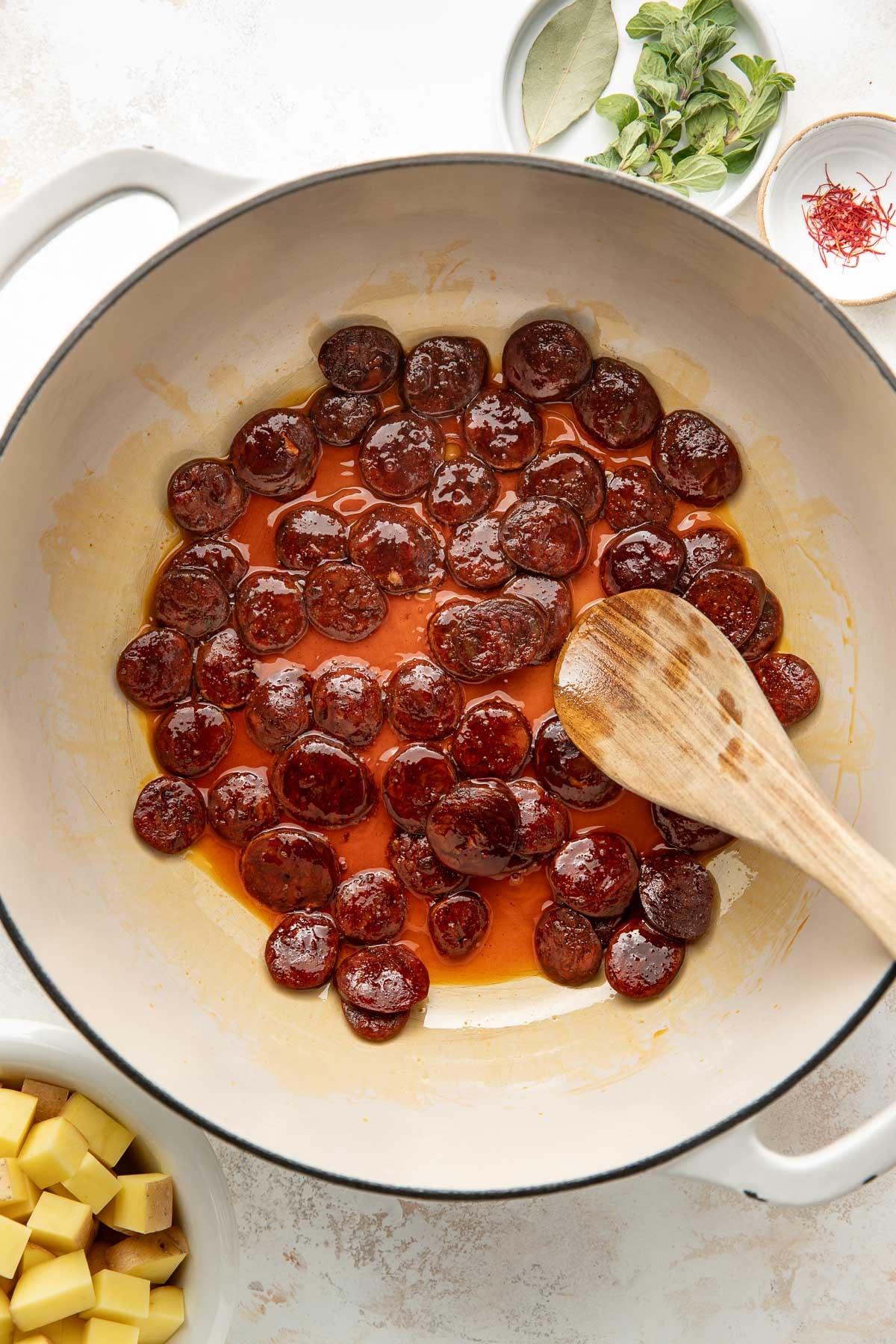 A white pot with sliced chorizo being sautéed, releasing red oil, stirred with a wooden spoon. Around the pot are bowls of herbs, spices, and diced potatoes.