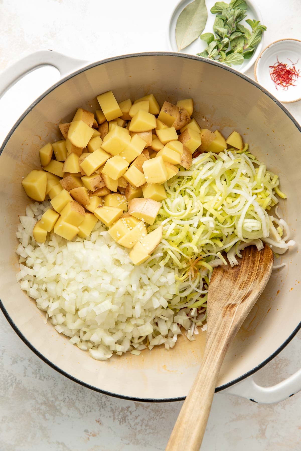 A white pot containing chopped potatoes, diced onions, and sliced leeks, with a wooden spoon resting inside. Small bowls with herbs and spices are visible in the background.