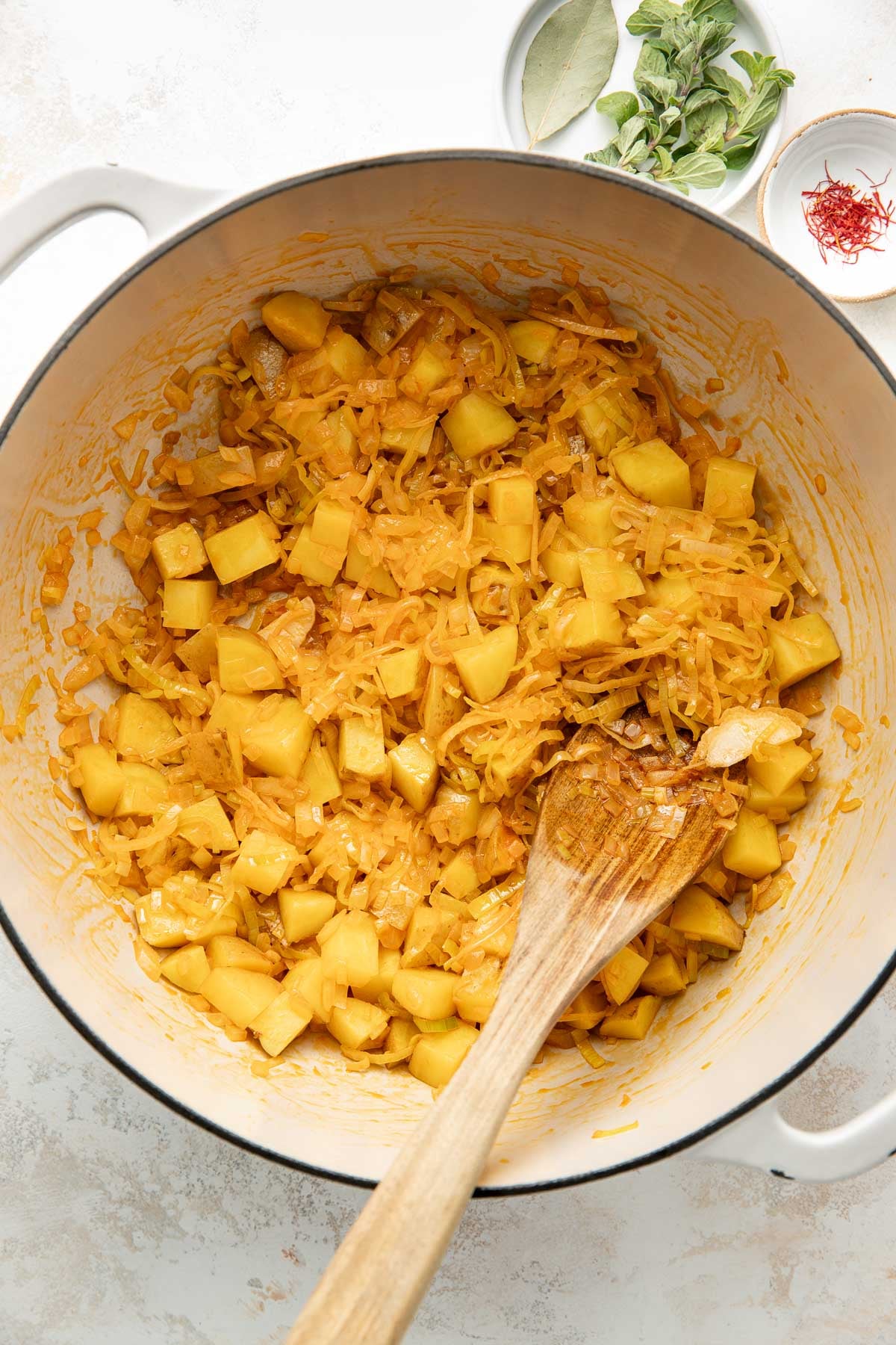 A white pot filled with diced potatoes and shredded onions cooked with spices. A wooden spoon is resting inside the pot. Nearby, small bowls hold bay leaves, fresh herbs, and saffron threads on a light surface.
