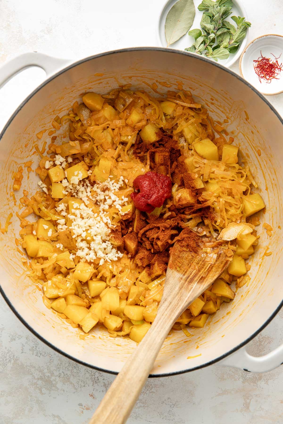 A white pot filled with chopped potatoes, grated onion, minced garlic, tomato paste, and spices being mixed with a wooden spoon. Bowls with bay leaves, fresh herbs, and saffron are beside the pot.