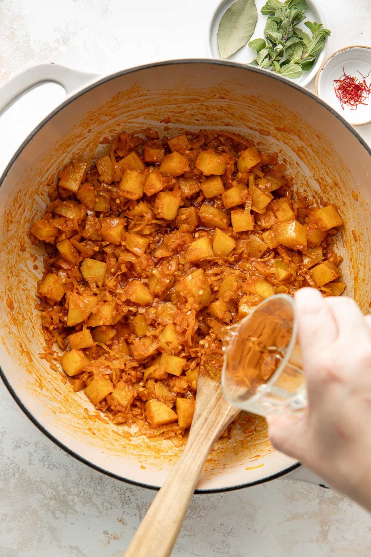 A hand pours liquid from a small glass cup into a pot containing diced potatoes, onions, and spices, being stirred with a wooden spoon. Bay leaves, oregano, and saffron are in small bowls nearby.