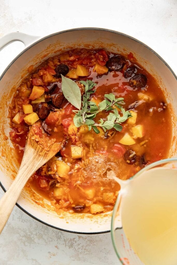 A pot of stew with tomato sauce, chopped potatoes, prunes, and fresh herbs, being stirred with a wooden spoon. A glass measuring cup of broth is being poured in from the side.