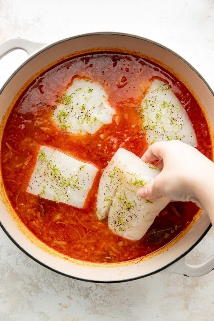 A hand is placing a piece of raw white fish fillet, seasoned with herbs, into a pot of red tomato-based sauce with other fillets. The pot is on a light-colored surface.