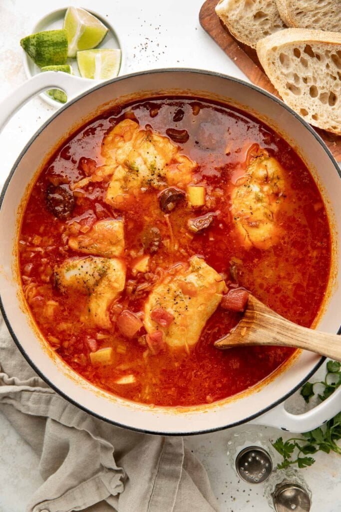 A pot of fish stew with chunky pieces of white fish in a red tomato-based broth, garnished with herbs. A wooden spoon rests in the pot. Sliced bread, lime wedges, and a beige napkin are nearby.