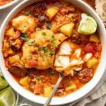 A bowl of fish stew with chunks of white fish, potatoes, tomatoes, and sausage, garnished with chopped herbs. A spoon rests in the bowl, and lime wedges and bread are visible nearby.