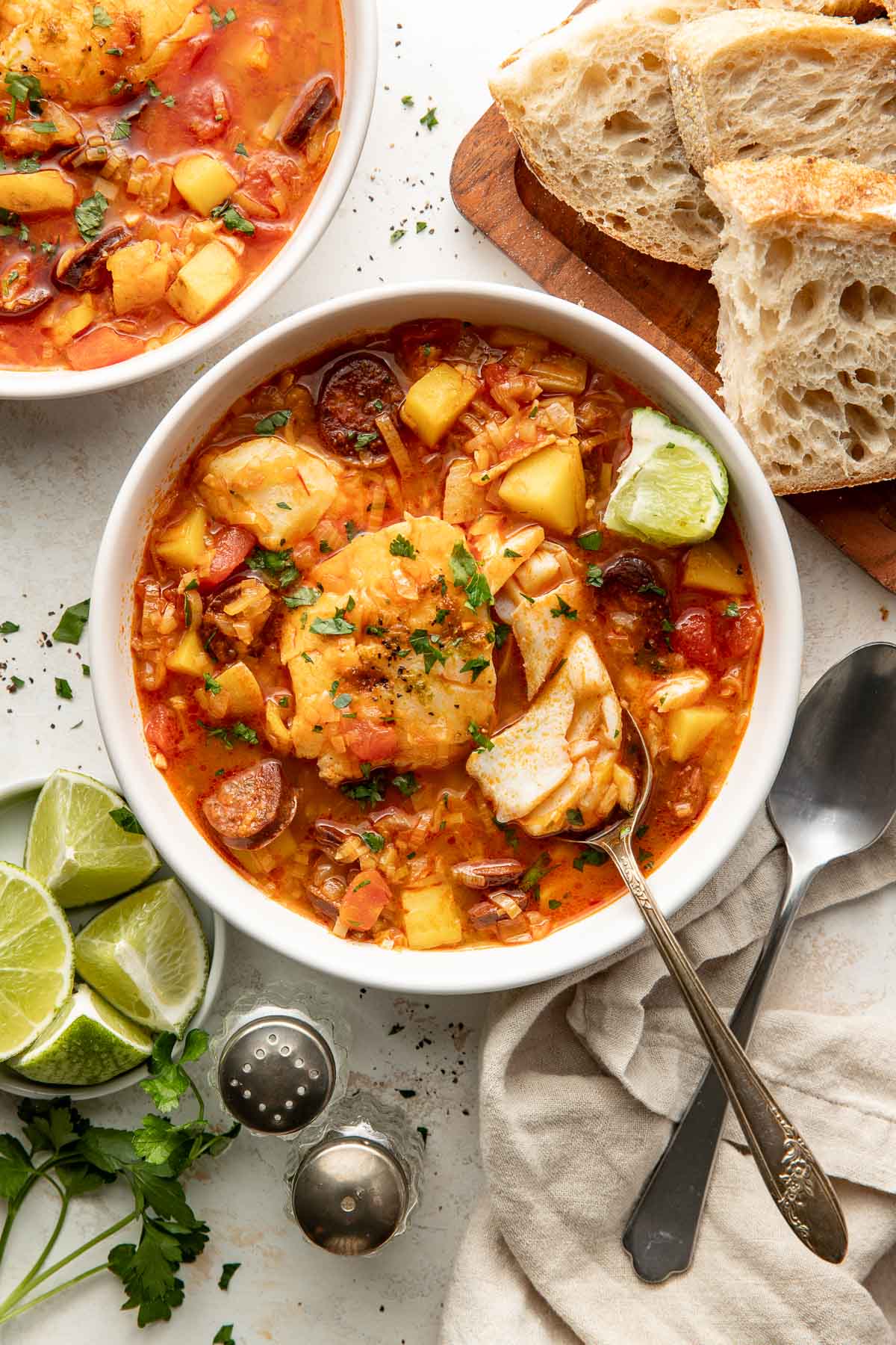 A bowl of fish stew with potatoes, tomatoes, and herbs, garnished with chopped parsley. A spoon rests in the bowl. Slices of bread, lime wedges, and parsley are nearby on the table.