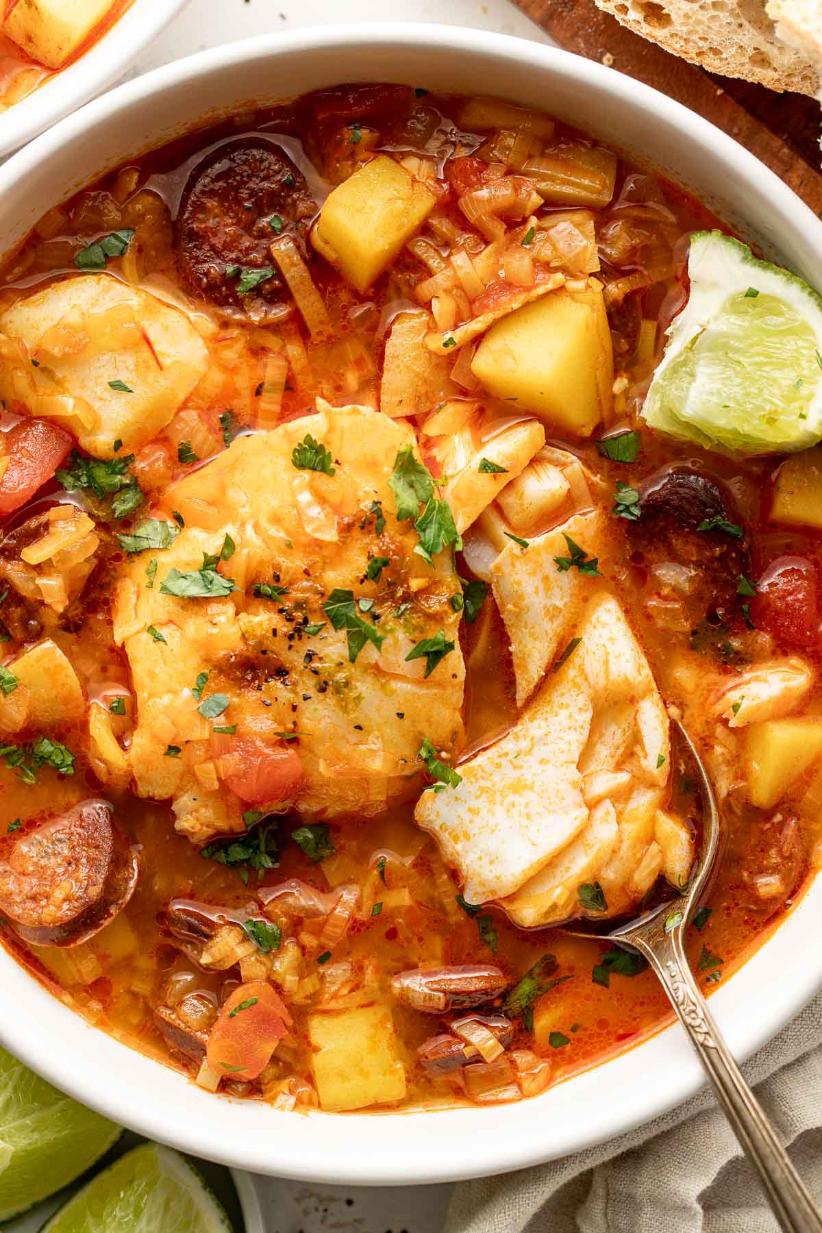 A bowl of fish stew with chunks of fish, potatoes, sausage, tomatoes, and herbs, garnished with chopped parsley and served with a wedge of lime and a spoon.