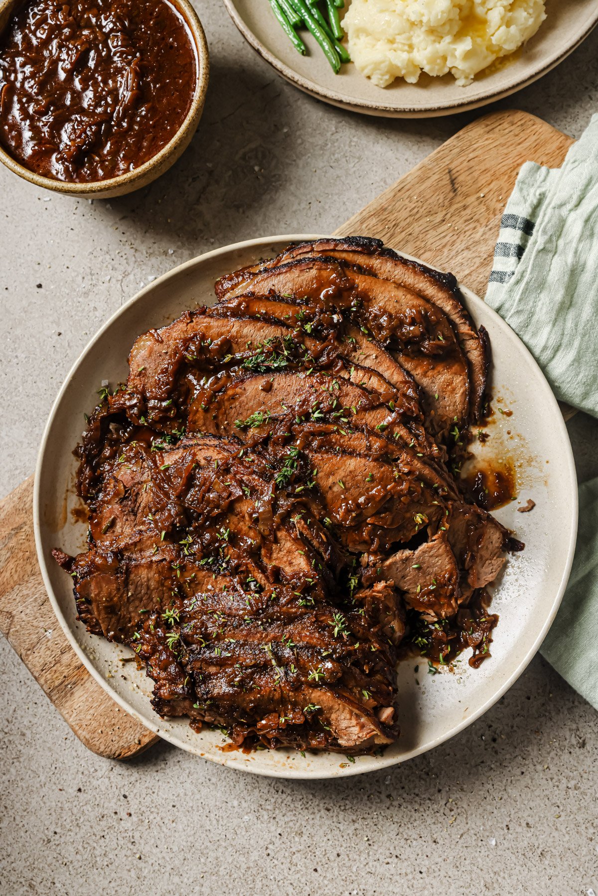 A plate of sliced, cooked beef brisket garnished with herbs sits on a wooden board. Nearby are mashed potatoes, green beans, and a bowl of sauce. The setting is rustic and inviting.