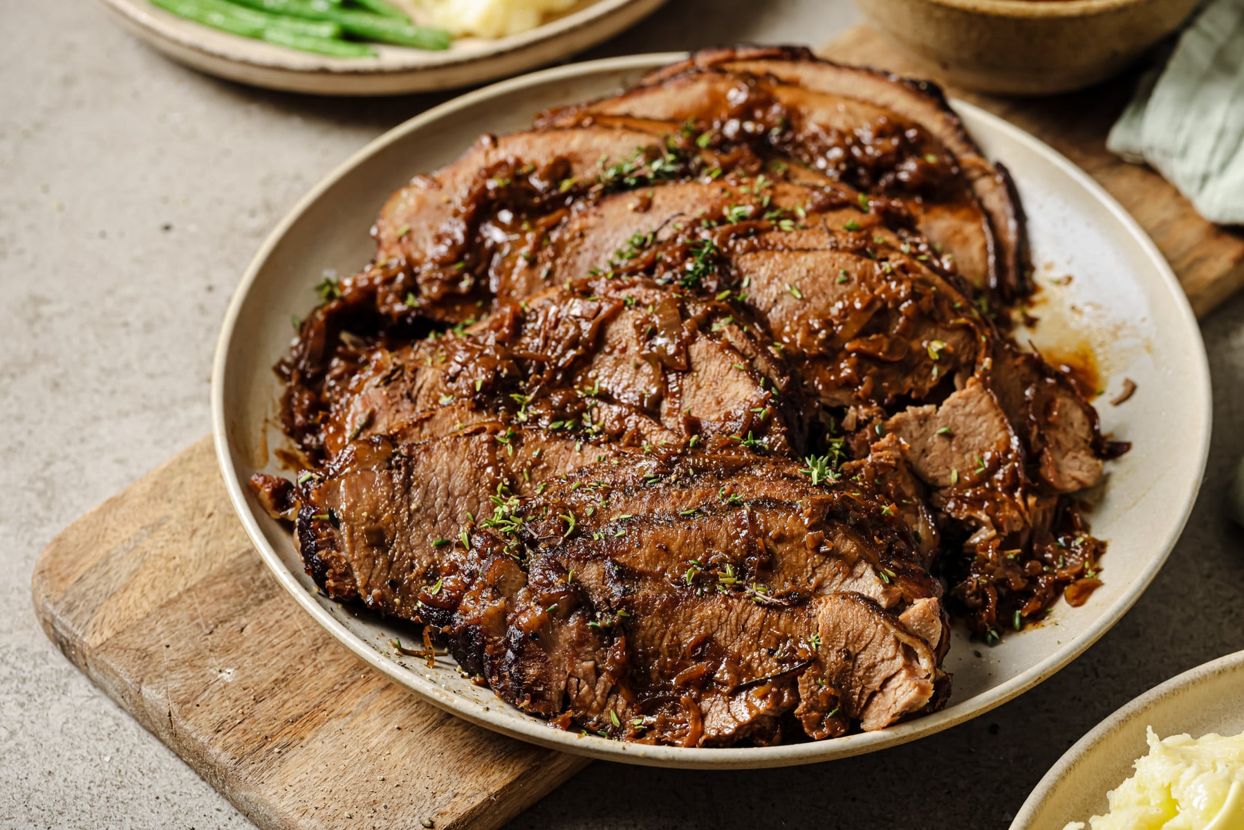 Sliced roast beef topped with herbs and a rich brown sauce, served on a round plate set on a wooden board, with sides of green beans and mashed potatoes in the background.