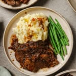 A plate with sliced pot roast topped with brown gravy, served alongside mashed potatoes with gravy and a portion of green beans. Other similar plates are partially visible around the main plate.