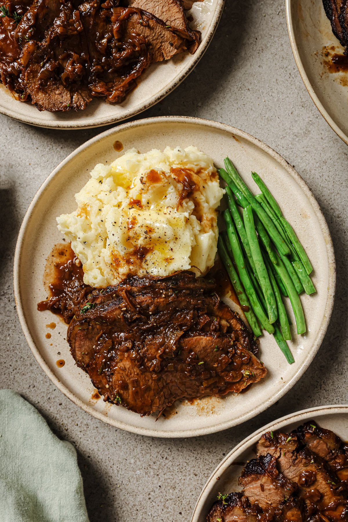 A plate with sliced pot roast topped with brown gravy, served alongside mashed potatoes with gravy and a portion of green beans. Other similar plates are partially visible around the main plate.
