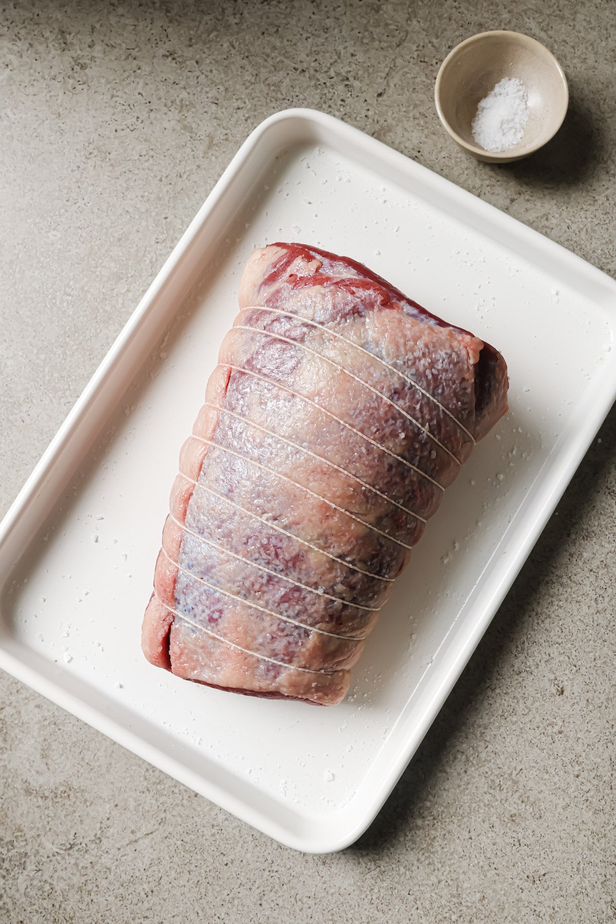 A raw, tied beef roast sprinkled with salt sits on a white tray. A small bowl of salt is nearby on a light-colored countertop.