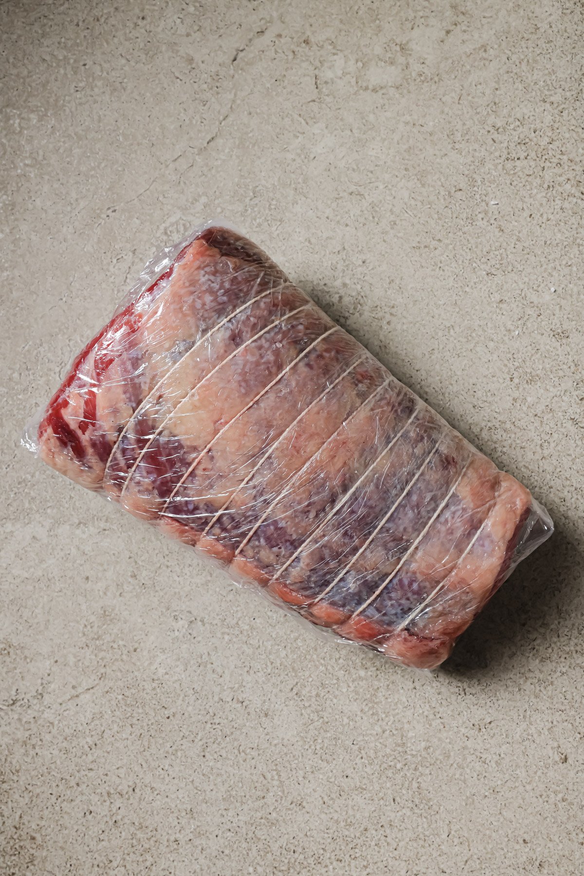 A raw beef roast wrapped in plastic and tied with butcher’s twine sits on a beige, textured surface.