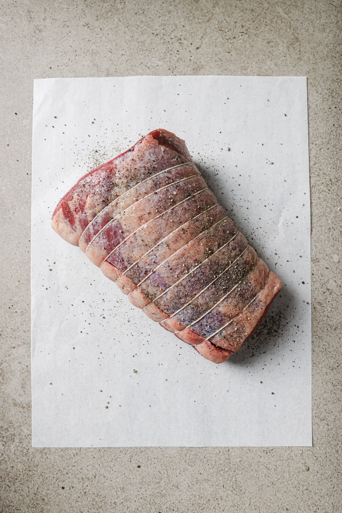 A raw, seasoned beef roast tied with butcher’s twine sits on a sheet of white parchment paper atop a light gray countertop. Black pepper and spices are visible on the meat and parchment.