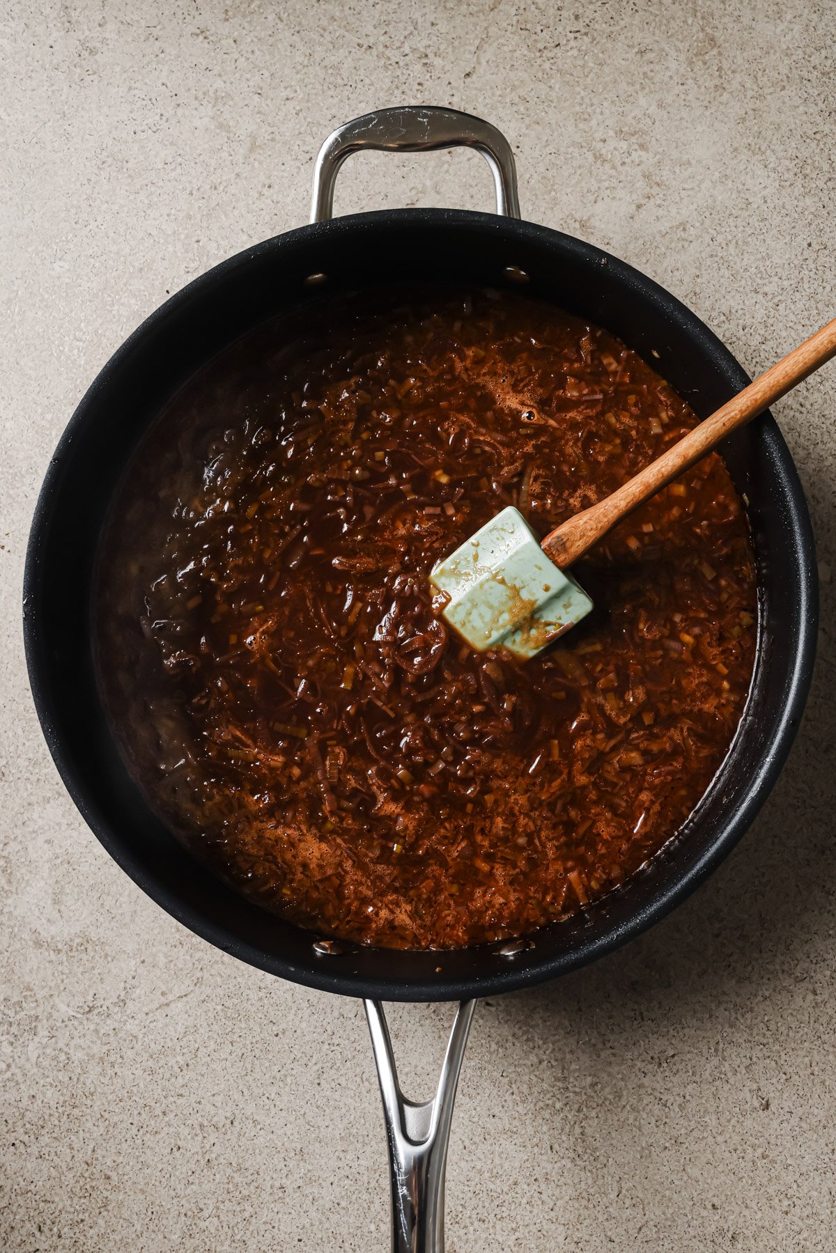 A black skillet filled with brown sauce and finely chopped onions sits on a light countertop. A blue spatula with a wooden handle rests in the sauce.