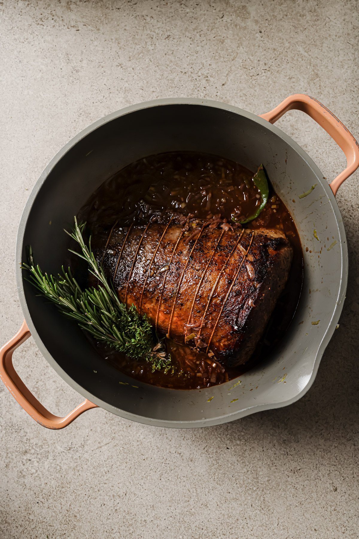 A browned roast tied with string sits in a round pot with handles, simmering in a rich brown sauce with herbs like rosemary, thyme, and a bay leaf on a light textured surface.