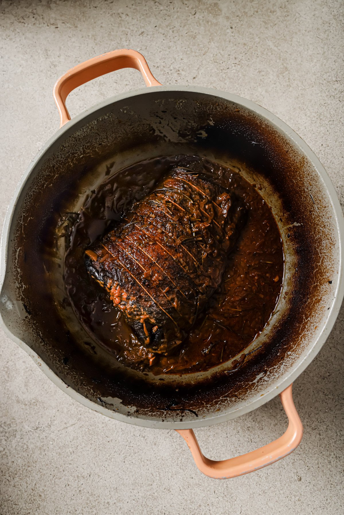 A browned, tied roast sits in a round, enameled Dutch oven filled with dark, rich sauce. The pot has orange handles and is placed on a light-colored surface.