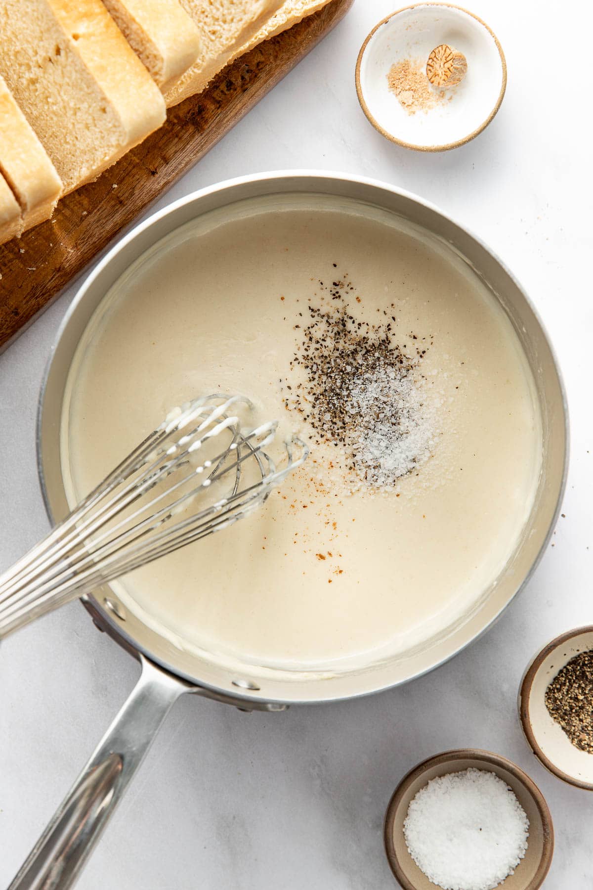 A saucepan filled with creamy white sauce being whisked, topped with salt, pepper, and grated nutmeg. Sliced bread sits on a cutting board, and nearby dishes hold spices.