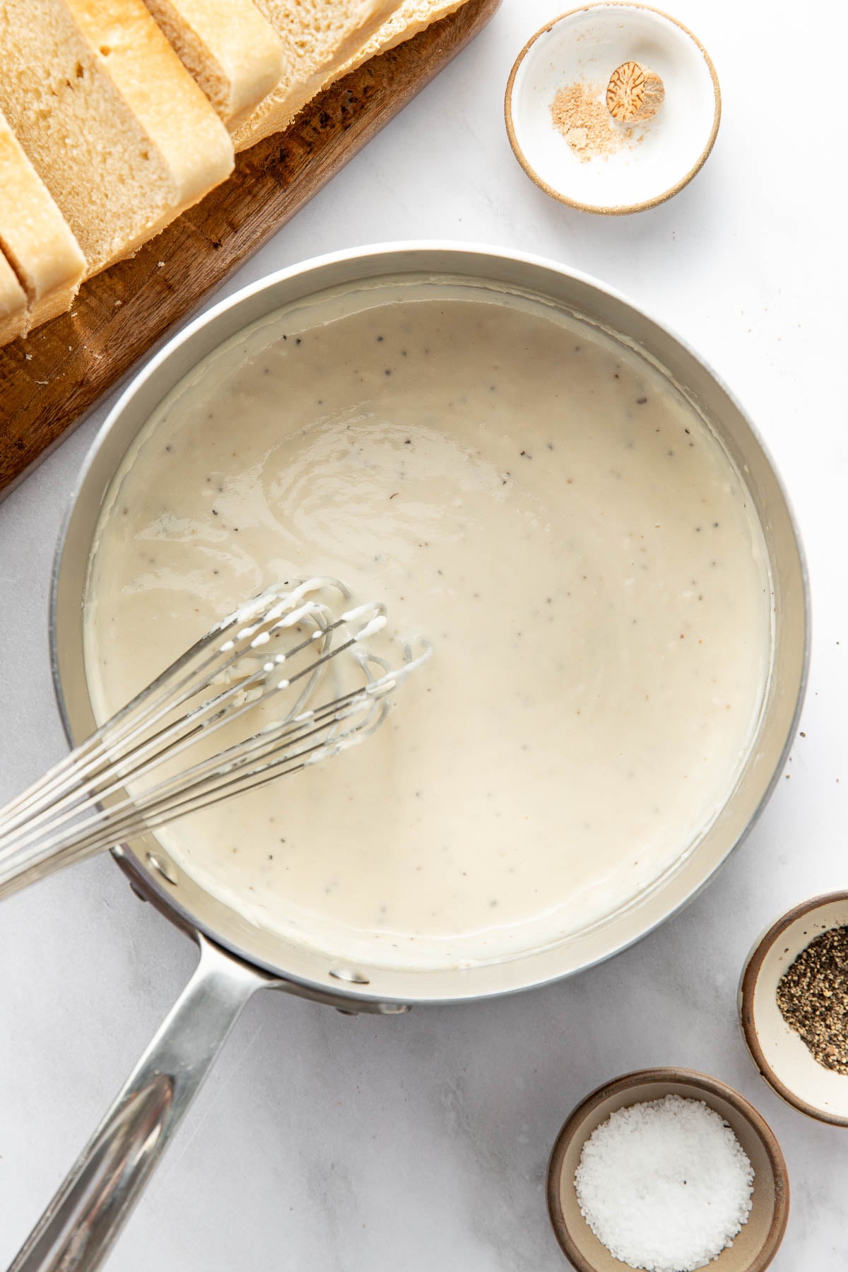 A saucepan filled with creamy white sauce being whisked, surrounded by bowls of salt, pepper, ground nutmeg, and slices of bread on a wooden board.