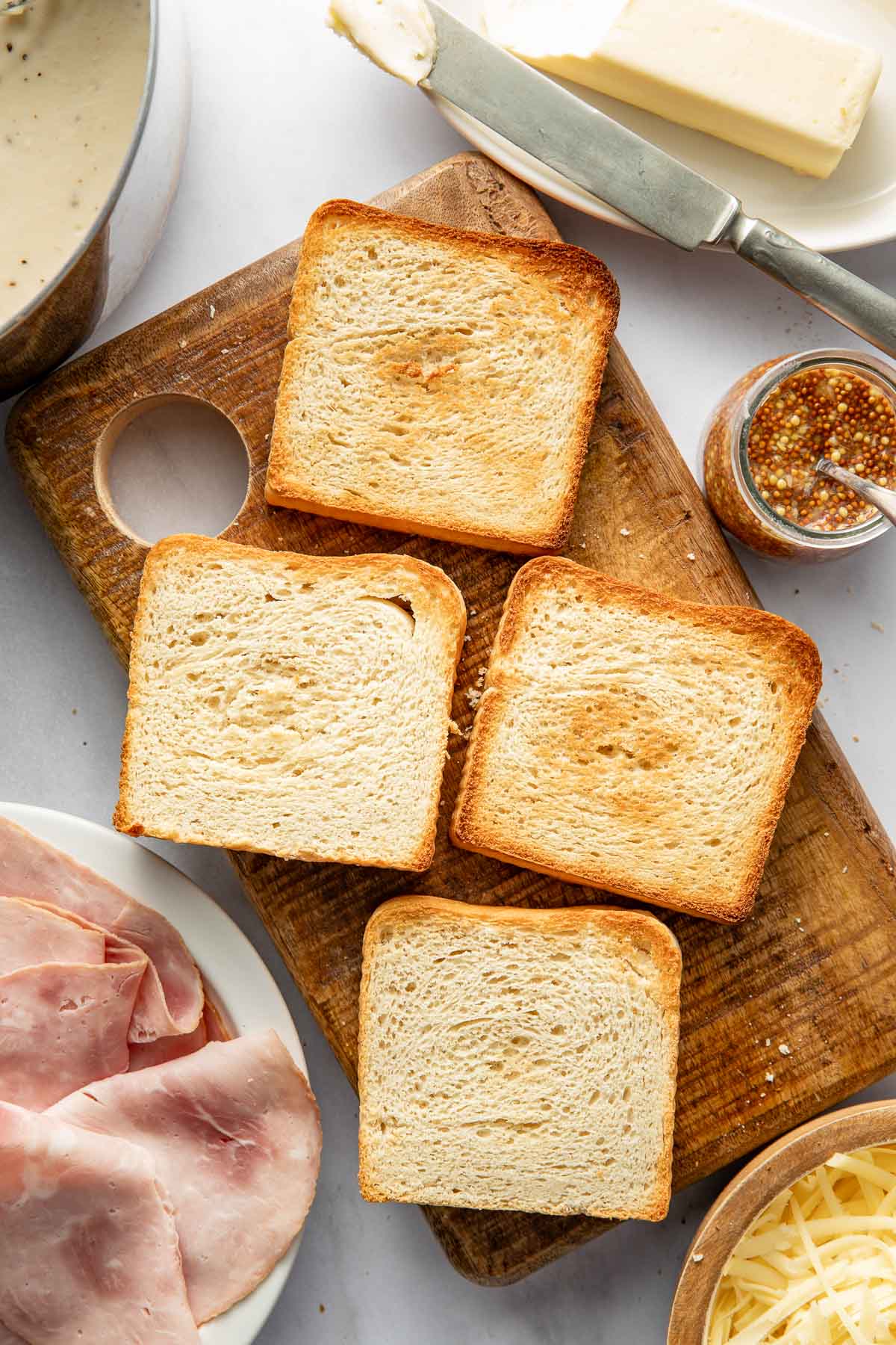 Four slices of toasted bread on a wooden cutting board, surrounded by a plate of sliced ham, a dish of grated cheese, whole grain mustard, and a stick of butter with a butter knife.