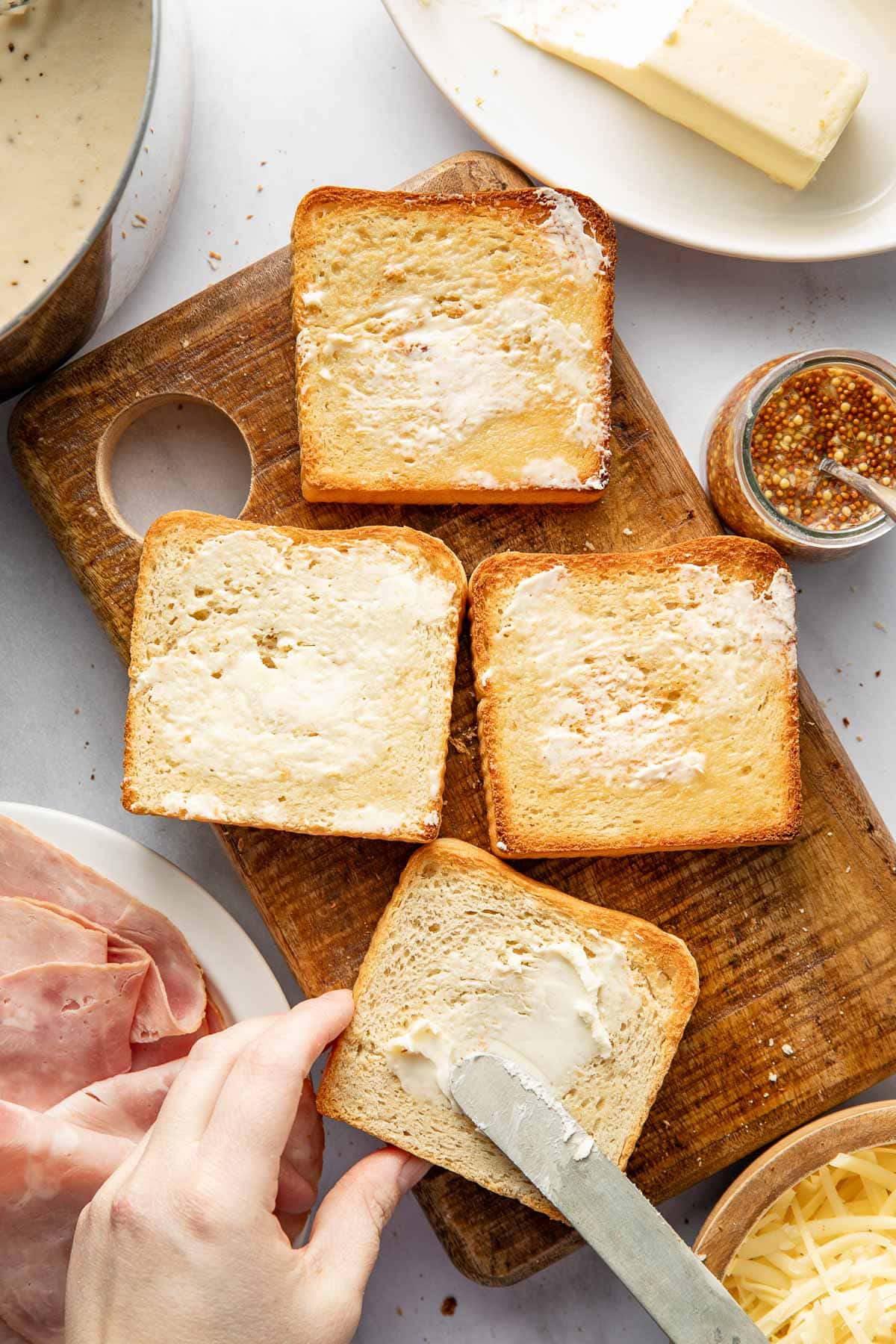 Four slices of toasted bread on a wooden board, three spread with butter. A hand is buttering one slice. Surrounding items include ham, shredded cheese, mustard, a stick of butter, and sauce.