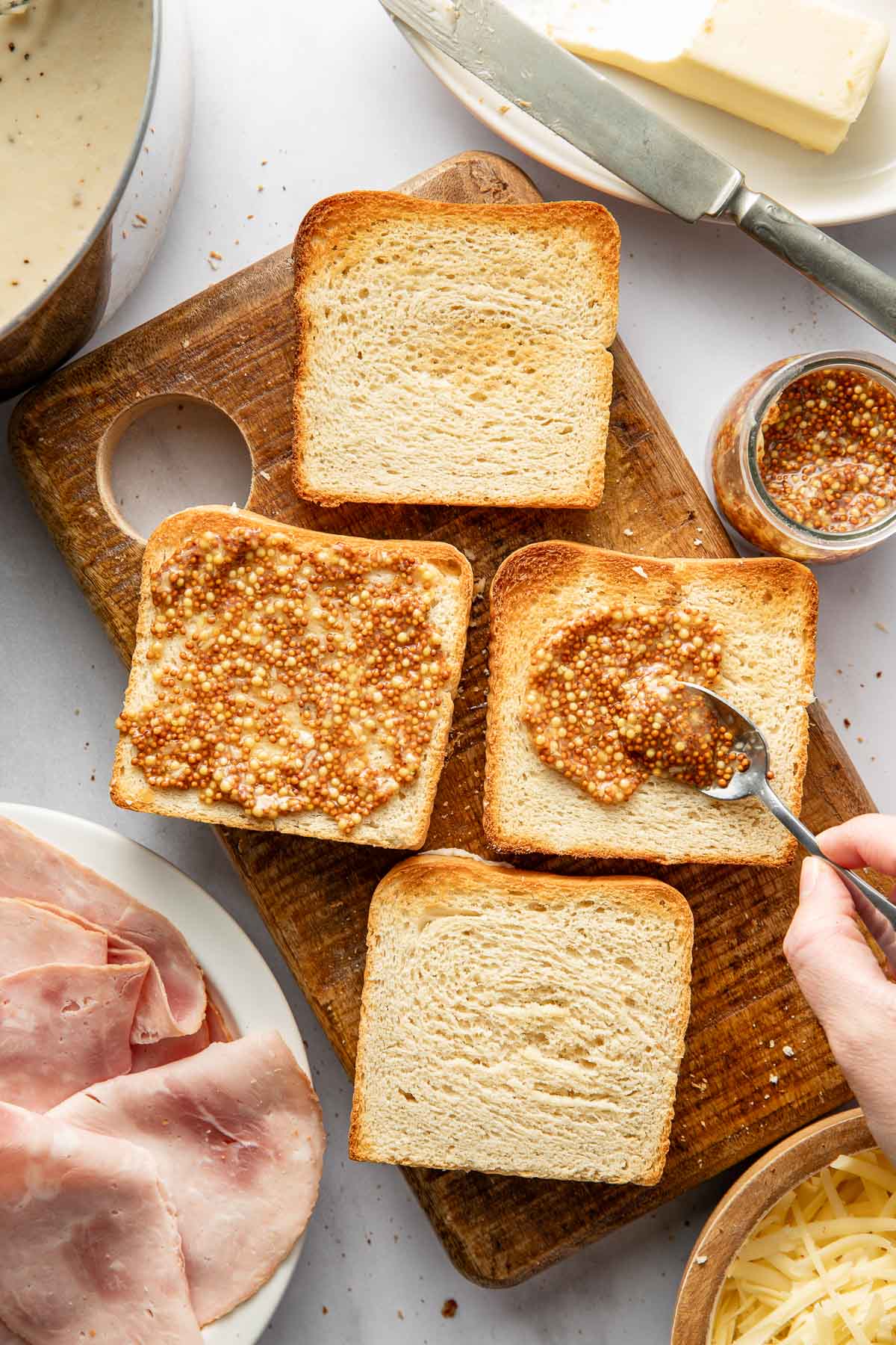 Four slices of bread on a wooden cutting board, with one being spread with grainy mustard. Nearby are a plate of ham, a bowl of shredded cheese, a jar of mustard, a knife, and a dish of butter.