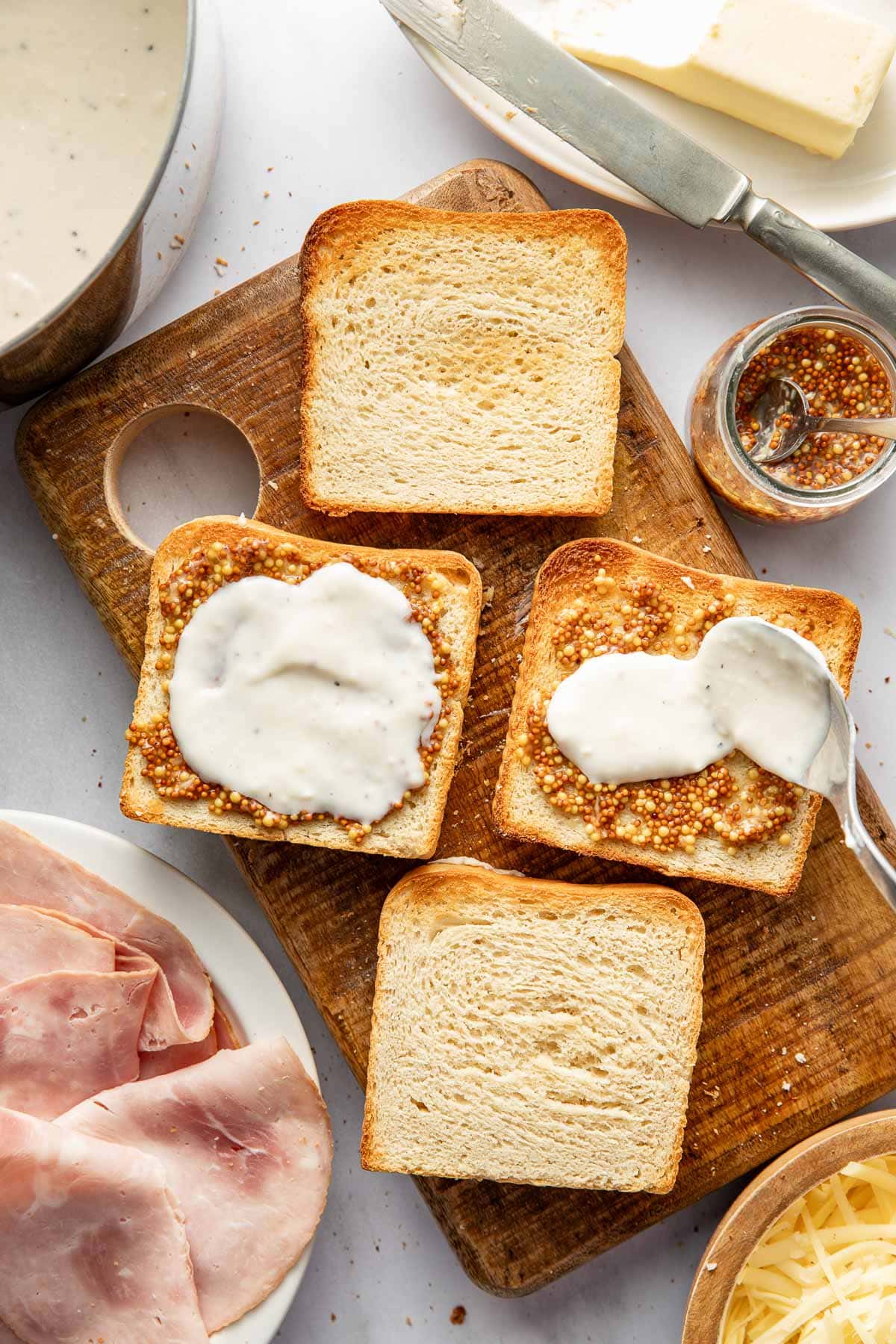 Four slices of bread on a wooden board, two topped with wholegrain mustard and creamy white sauce. Slices of ham, shredded cheese, butter, and a jar of mustard are nearby. A spoon spreads sauce on one slice.