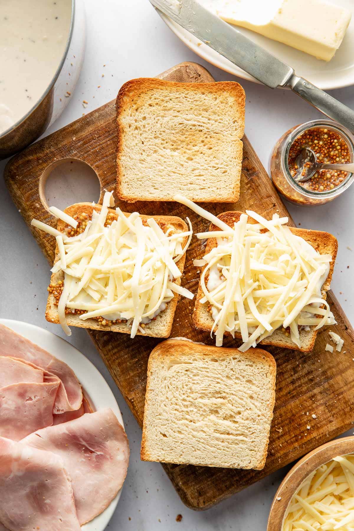 Three slices of bread topped with grated cheese sit on a wooden cutting board. Surrounding them are slices of ham, butter, grainy mustard, a bowl of white sauce, and more grated cheese.