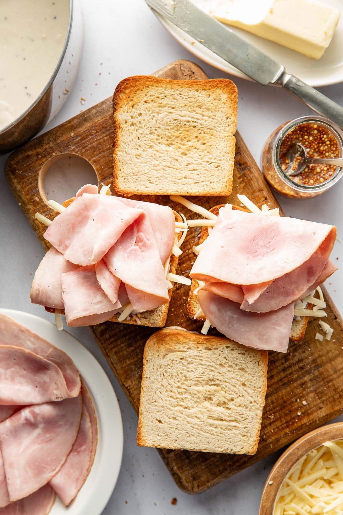 Slices of ham and shredded cheese are placed on slices of toasted bread on a wooden board, with mustard, butter, and extra ham and cheese nearby, ready to assemble sandwiches.