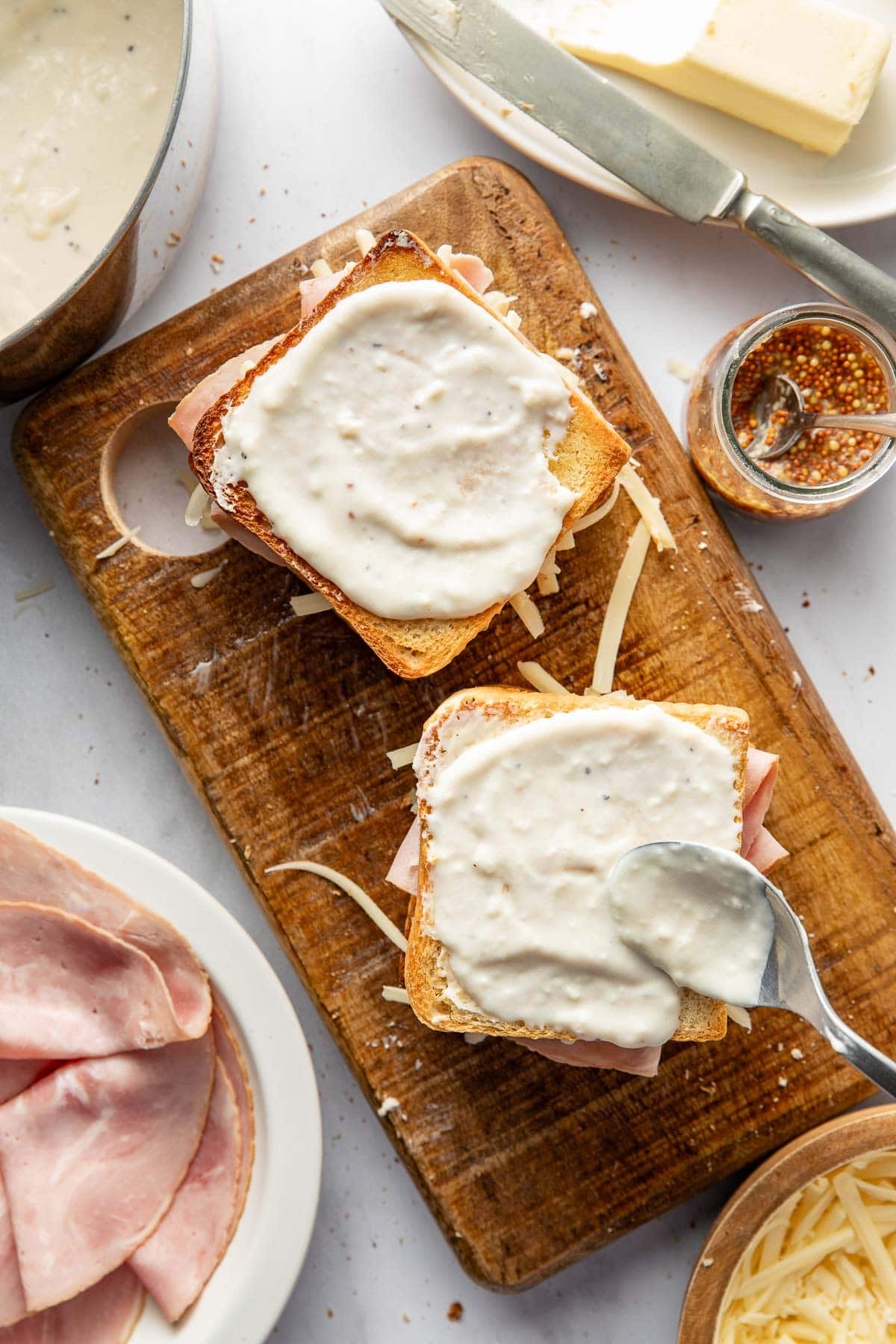 Two open-faced ham and cheese sandwiches on toasted bread sit on a wooden cutting board. A spoon spreads creamy white sauce on top. Nearby are ham slices, grated cheese, butter, mustard, and a bowl of sauce.