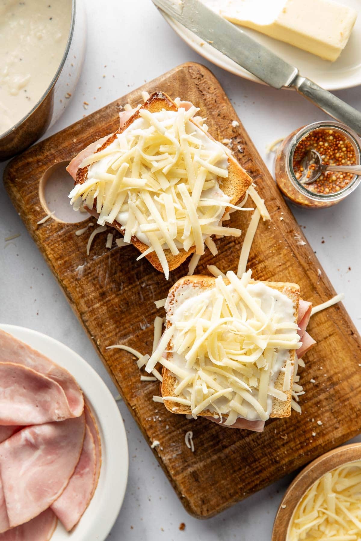 Two slices of bread topped with ham, grated cheese, and white sauce sit on a wooden cutting board. Nearby are a dish of ham slices, a jar of mustard, a knife with butter, and a pot of creamy sauce.