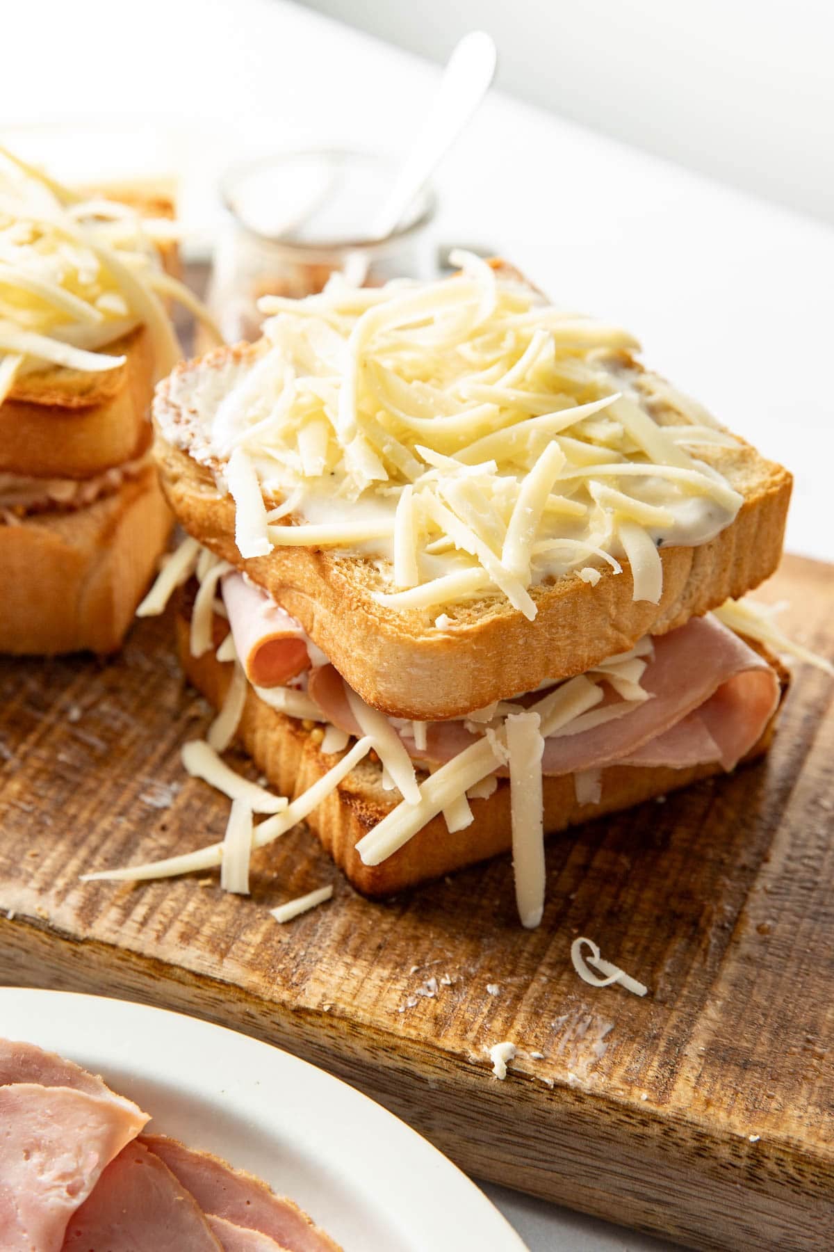 Two slices of bread layered with ham and shredded cheese, with extra cheese on top, sit on a wooden cutting board. A plate of ham and a small jar are visible in the background.