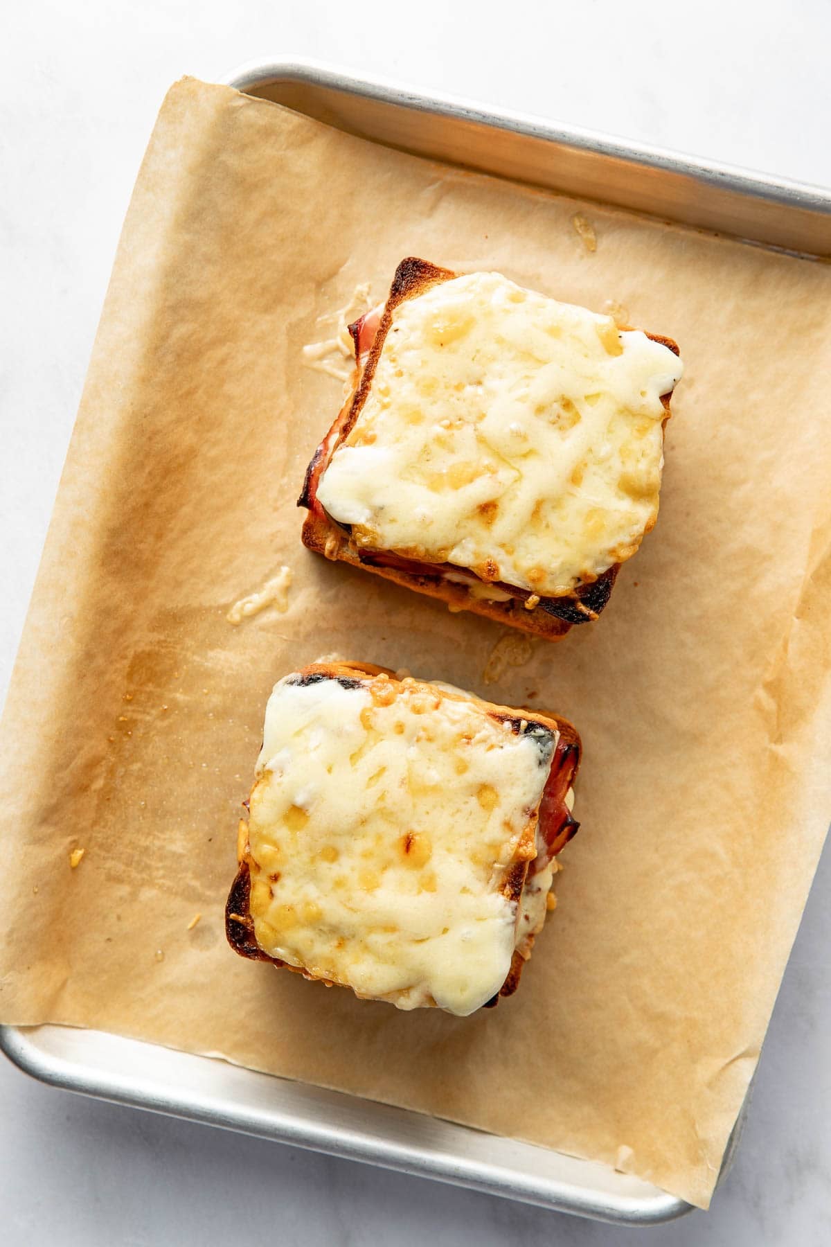 Two toasted sandwiches topped with melted cheese sit on a parchment-lined baking sheet. The sandwiches appear golden brown and slightly crisp around the edges.