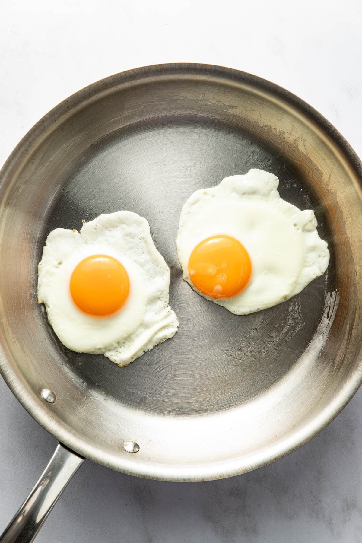 A stainless steel skillet containing two sunny-side up eggs with bright yellow yolks, cooked on a white marble surface.
