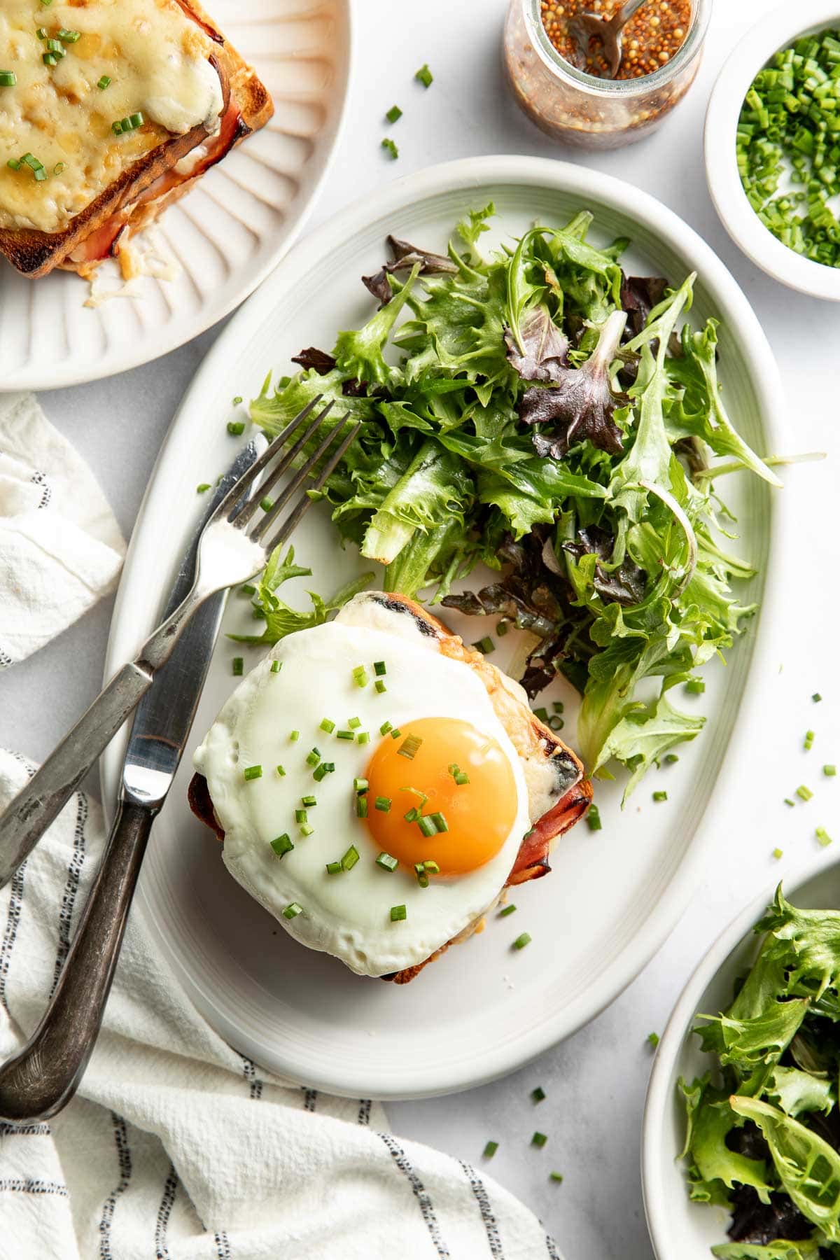 A plate with a Croque Madame topped with a sunny-side-up egg and chopped chives, served with a side of mixed green salad. A fork and knife rest beside the plate, and extra greens, herbs, and condiments are nearby.