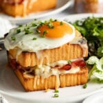 A Croque Madame sandwich topped with a sunny-side-up egg and chopped chives, filled with ham and melted cheese, served on a white plate with a side salad.